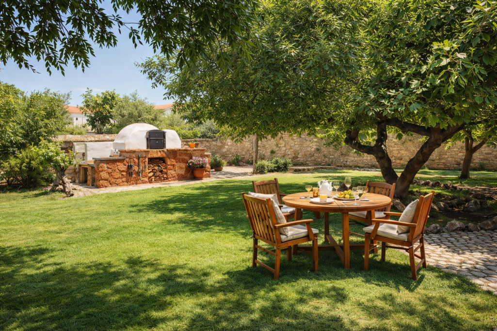 Mesa con sillas en un jardín, rodeada de árboles, con una cocina exterior al fondo.