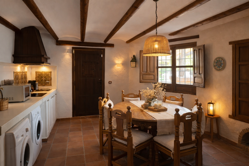 Cocina rústica con mesa, sillas de madera y decoración acogedora. Luz cálida y ambiente íntimo.