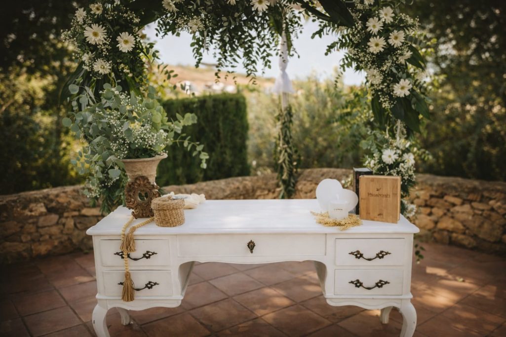Mesa blanca decorada con flores y elementos naturales en un entorno al aire libre.